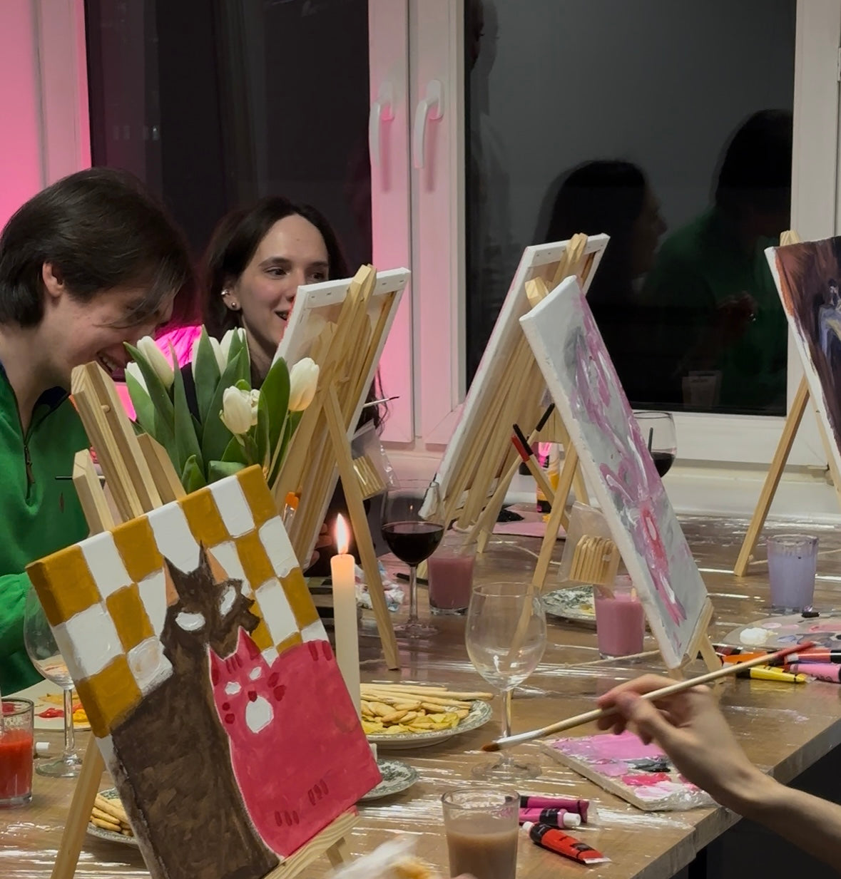 People painting at a table with easels and art supplies in a casual setting.